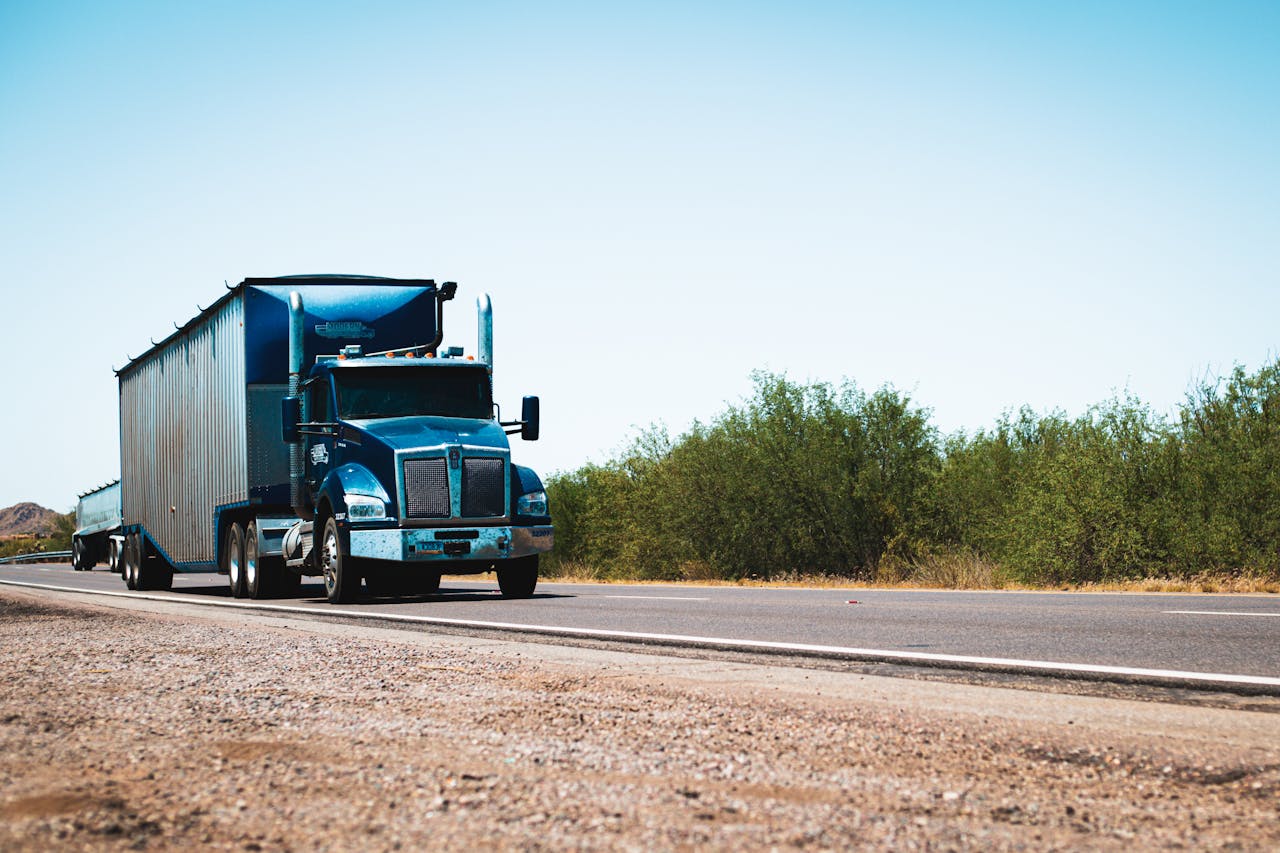 who-we-are A blue semi truck driving on a rural highway under clear blue skies, showcasing transportation and travel.