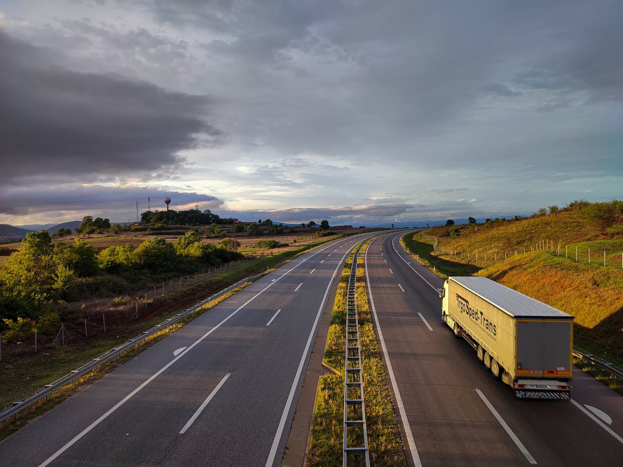 our-stories A scenic highway under a dramatic cloudy sky with a lone truck driving on an open road.